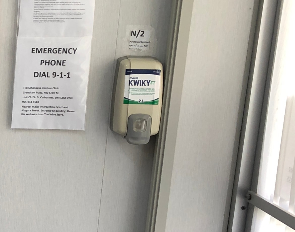 Antibacterial hand sanitizer dispenser on wall by the front door, next to signage for dialing 9-1-1 in an emergency and directions for the dispatcher