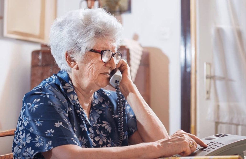 Elderly woman dialing a desktop phone with receiver against her ear