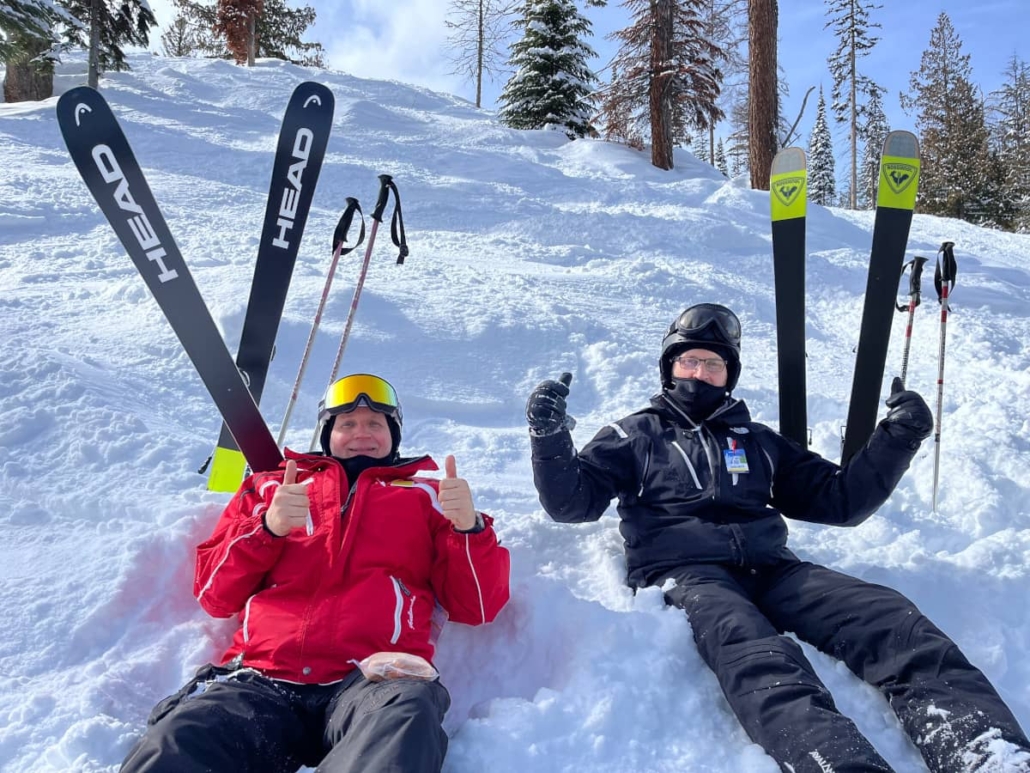 Your denturist Tim with thumbs up sitting on steep skiing slope having stopped for lunched with his skis and poles stuck upwards in snow, with one of his buddies next to him