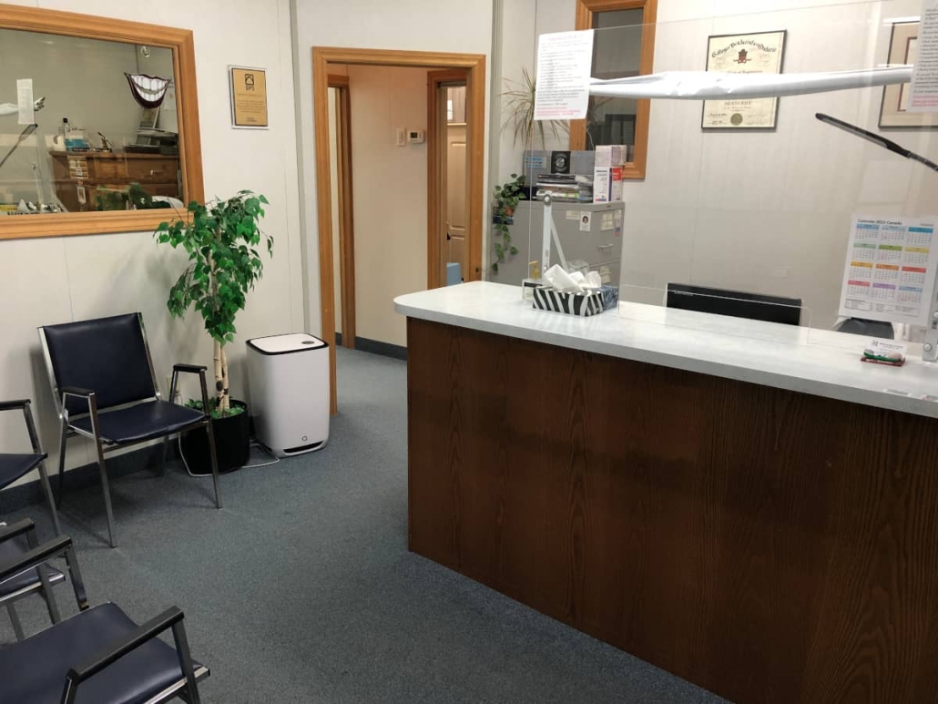 Wide shot of our reception desk, some waiting room chairs, some plants, a floor air purifier, large one-way mirror/glass window into the lab and a doorway leading to the rest of the clinic